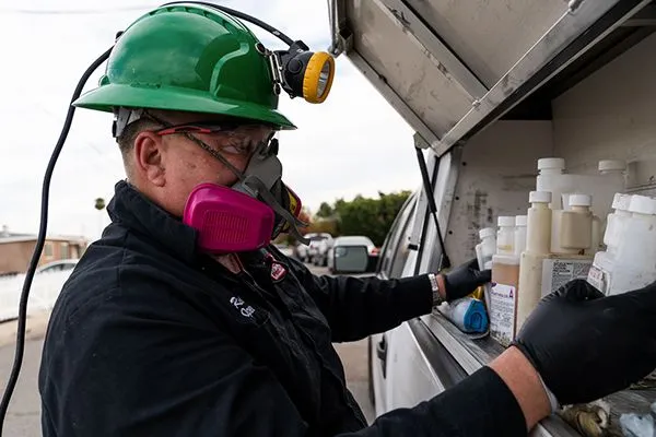 - Housecall Pro Pest control pro with hardhat and face mask gathers chemical bottles from work truck