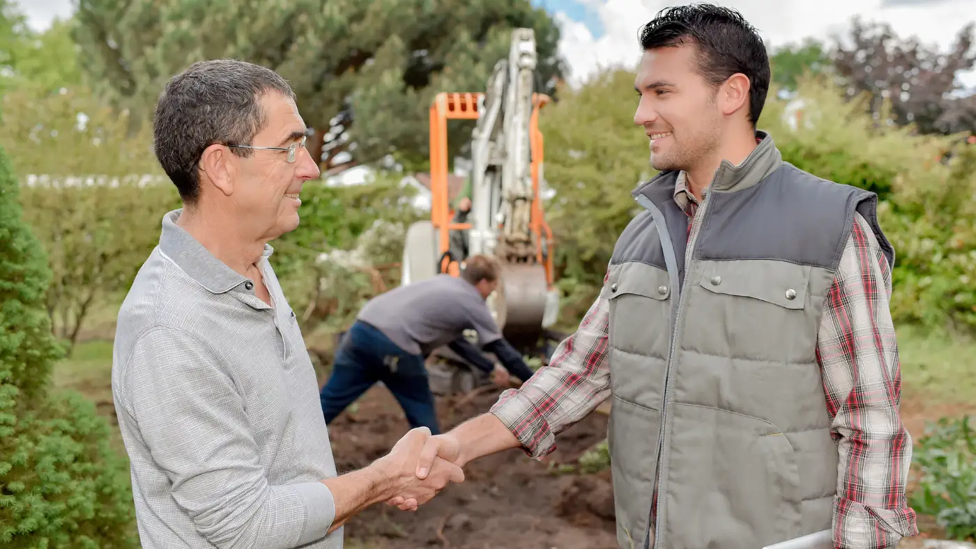 Landscaper shaking hands with a customer while holding a contract, with crew working in the background