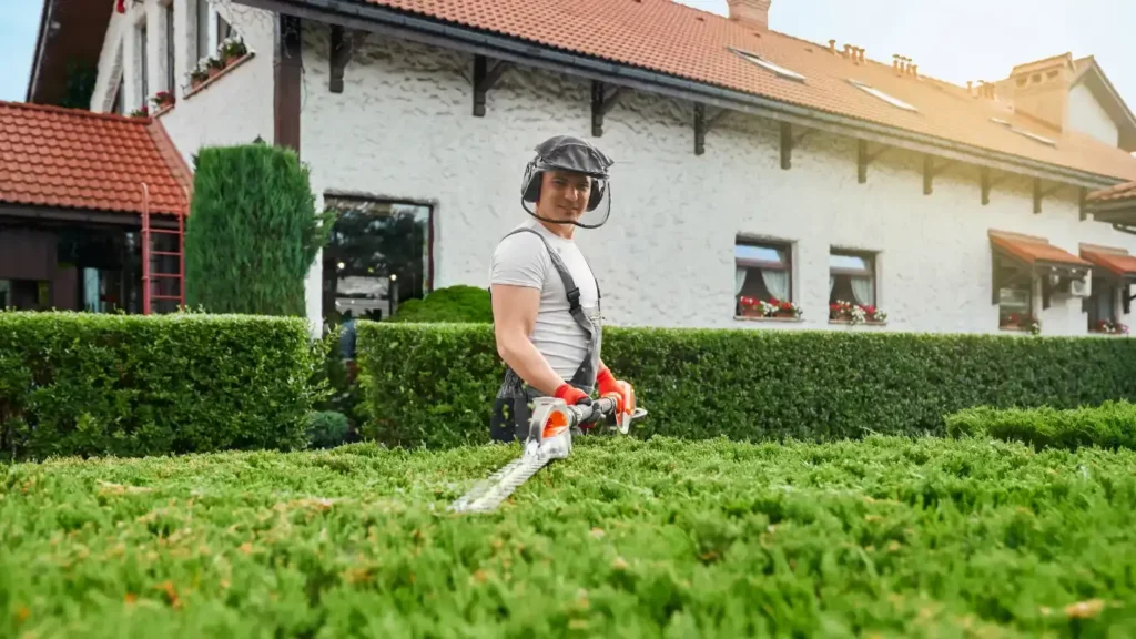 - Housecall Pro landscaping pro in uniform, glasses, and gloves pruning bushes in garden.