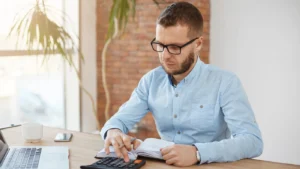 Small business owner calculating payroll expenses using a calculator and notebook at a desk.