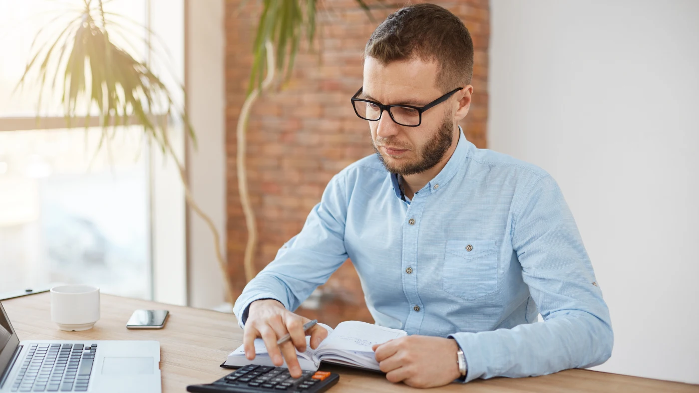 Small business owner calculating payroll expenses using a calculator and notebook at a desk.