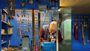 Experienced locksmith cutting a key at a workbench inside a shop lined with hundreds of keys and lock hardware.