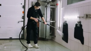 Worker using a pressure washer wand to clean tiled walls inside a commercial wash bay.